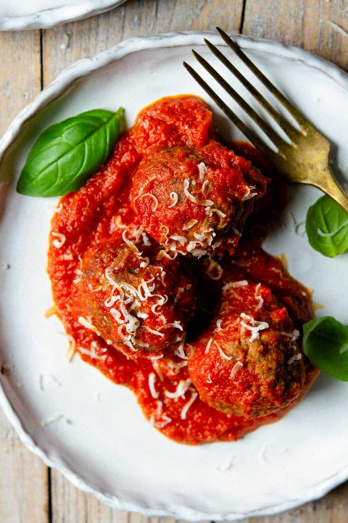 An overhead close up of three Italian meatballs on a plate with tomato sauce and basil leaves