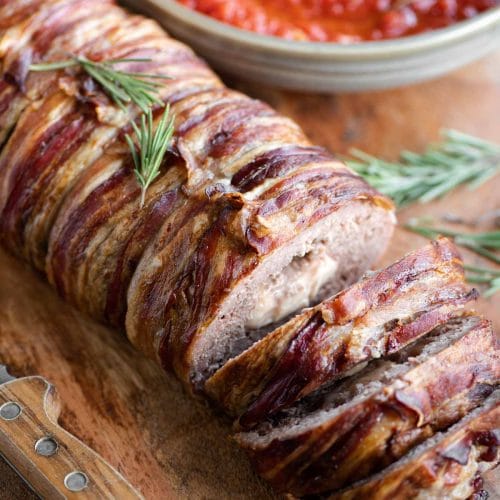 An Italian meatloaf wrapped in pancetta sitting on a wooden cutting board with some slices cut
