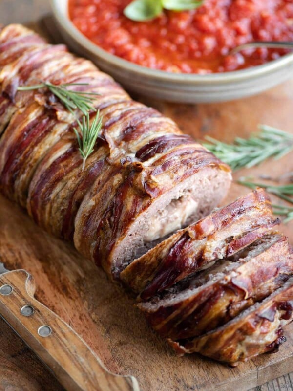 An Italian meatloaf wrapped in pancetta sitting on a wooden cutting board with some slices cut