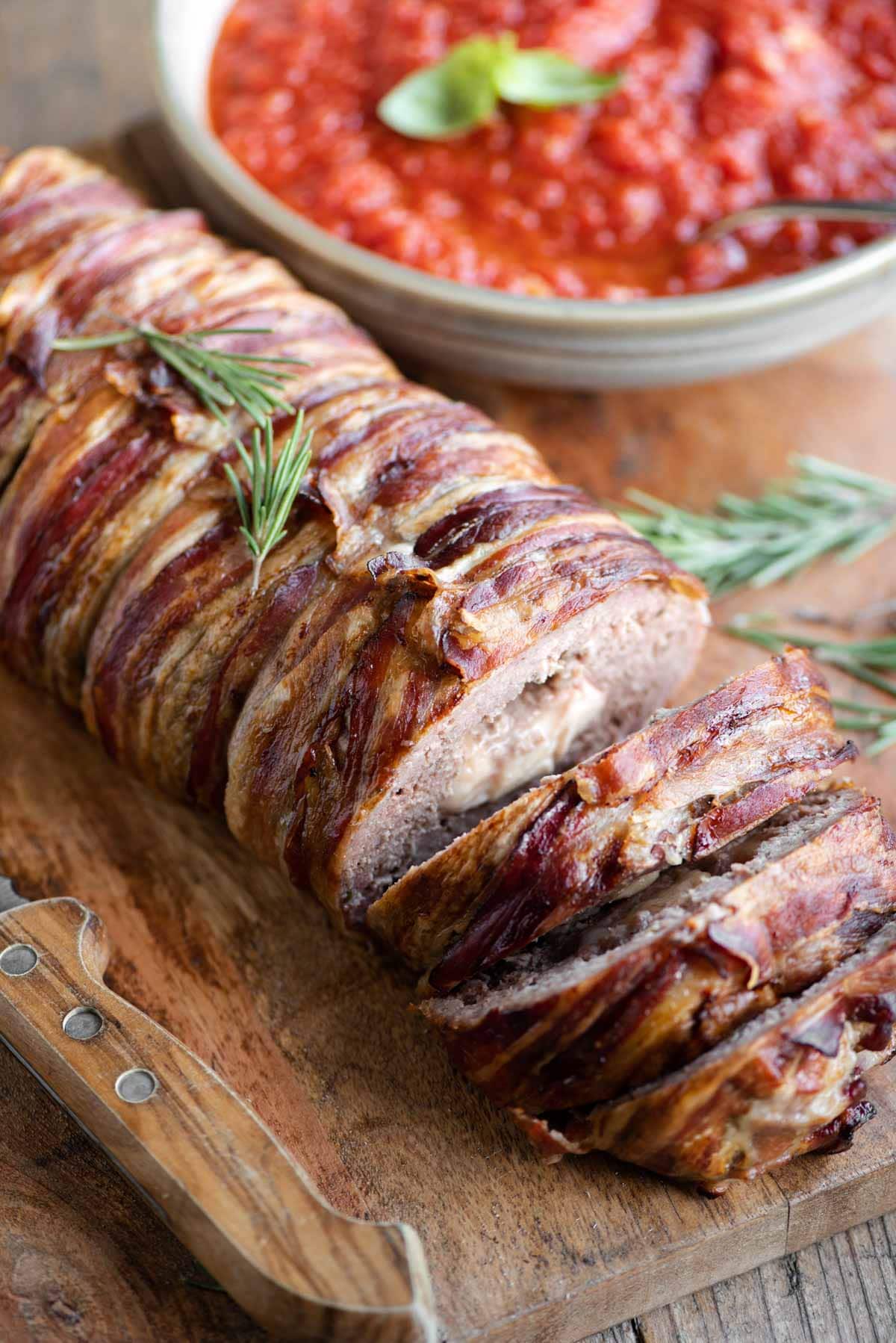 An Italian meatloaf wrapped in pancetta sitting on a wooden cutting board with some slices cut