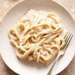 A close up of cacio e pepe with pici pasta on a plate with a fork