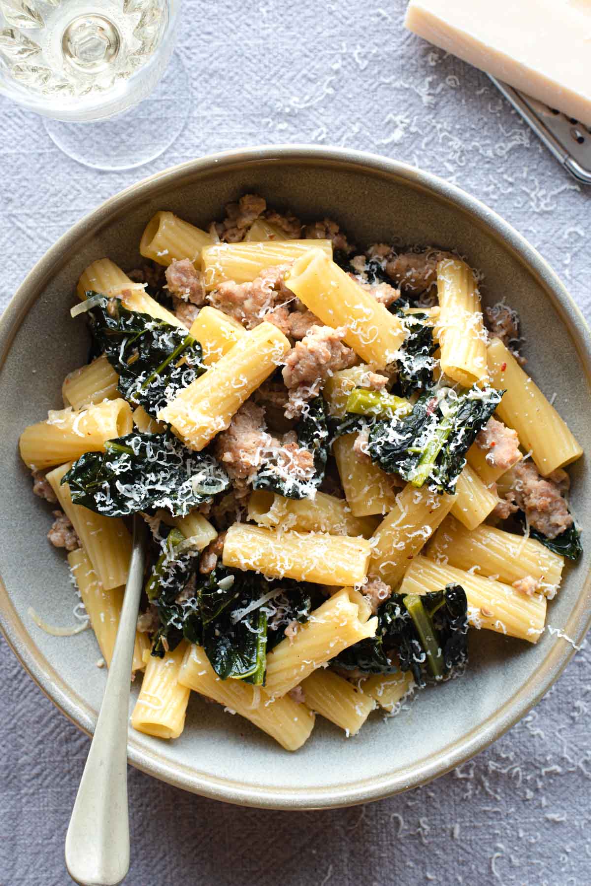 An overhead shot of a bowl of pasta with kale and sausage with a glass of wine at the side