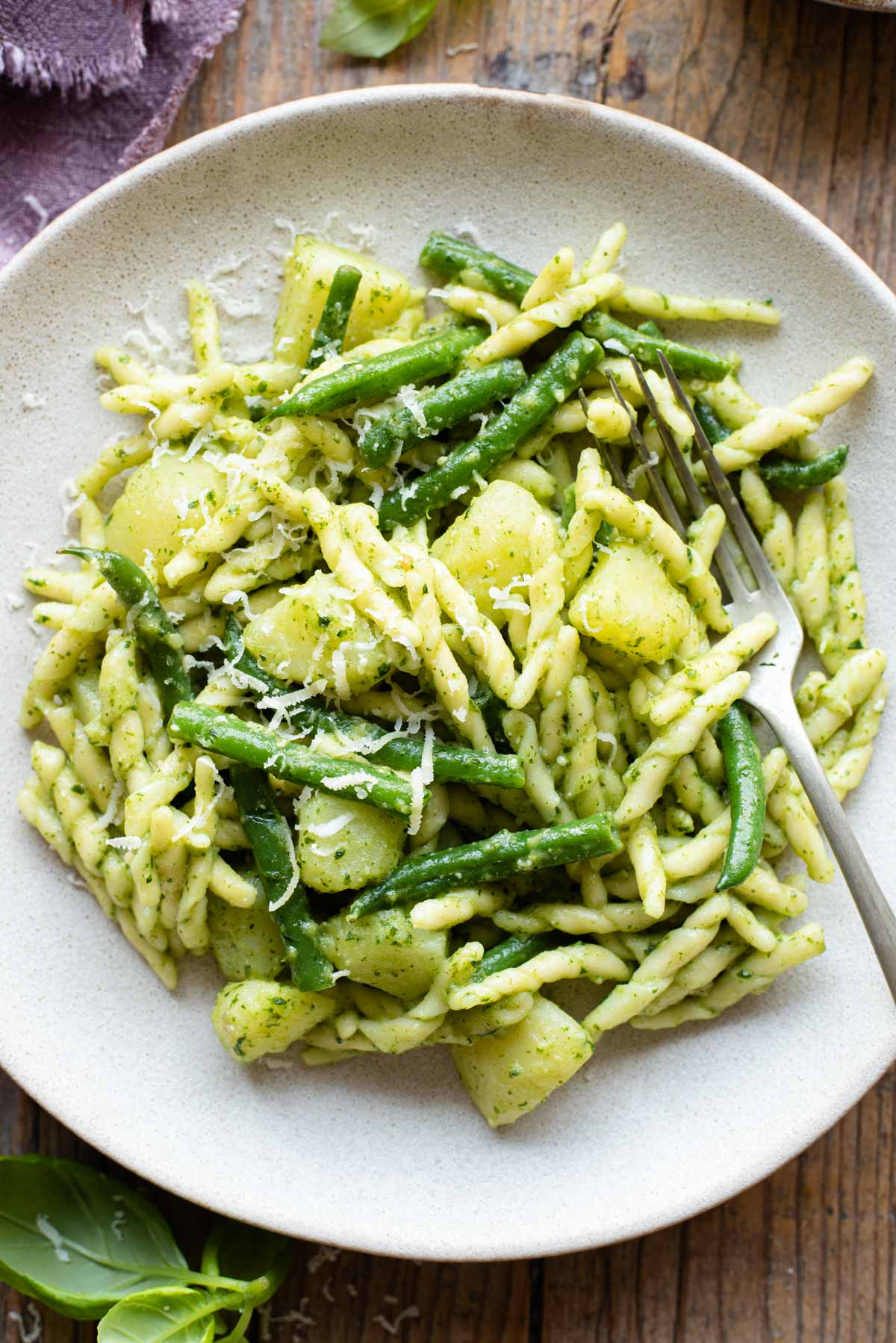 A close up of trofie pasta with pesto, potatoes and green beans on a plate with a fork at the side
