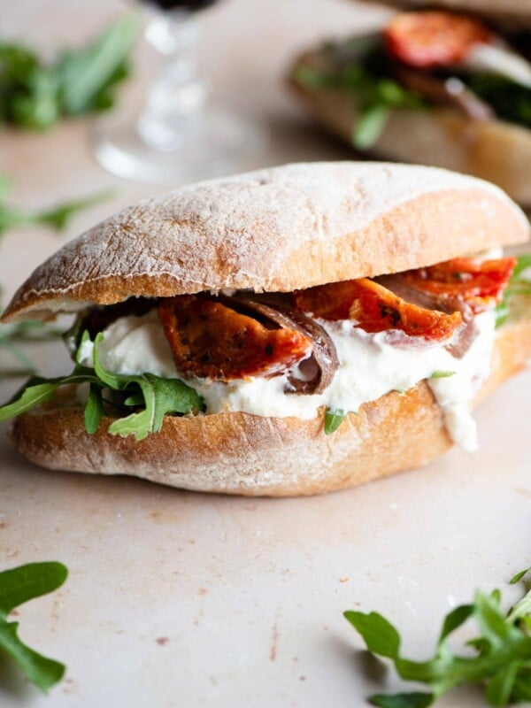 A side shot of a ciabatta sandwich with burrata, anchovies, sun blushed tomatoes and arugula on a light stone surface