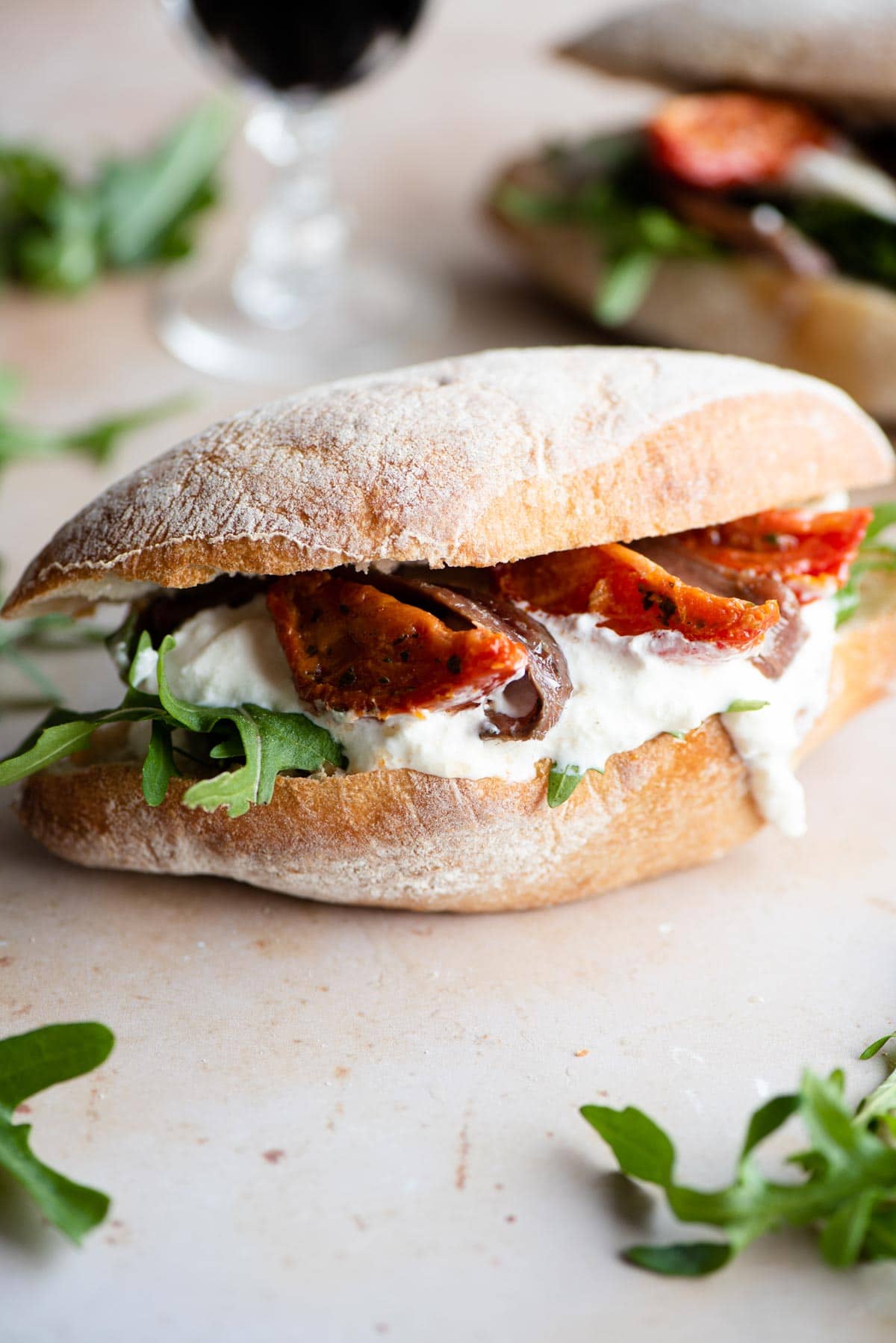 A side shot of a ciabatta sandwich with burrata, anchovies, sun blushed tomatoes and arugula on a light stone surface