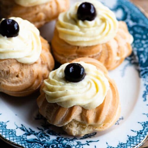 A close up of Italian Zeppolechoux buns filled with pastry cream and a cherry on top sitting on a blue and white plate.
