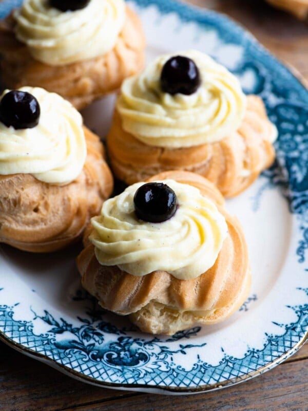 A close up of Italian Zeppolechoux buns filled with pastry cream and a cherry on top sitting on a blue and white plate.