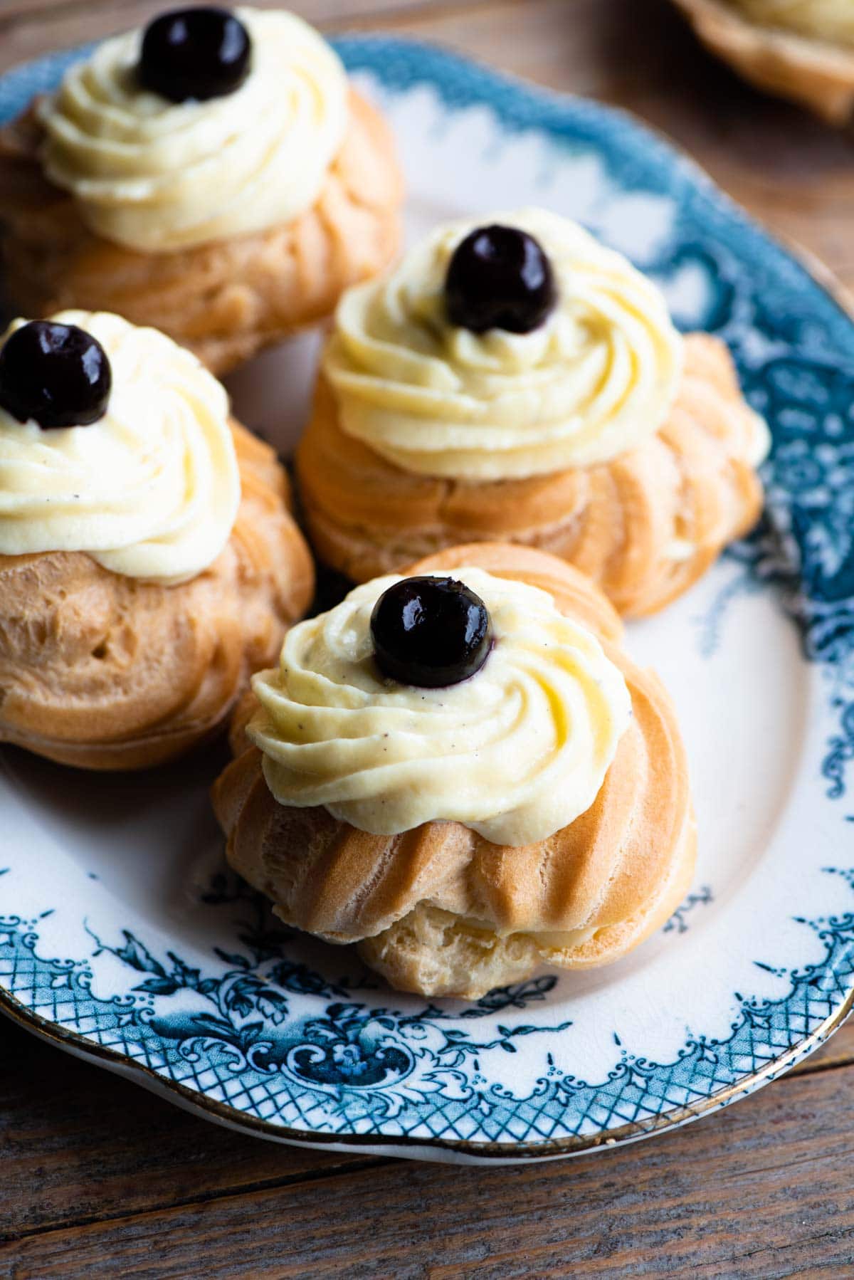 A close up of Italian Zeppole choux buns filled with pastry cream and a cherry on top sitting on a blue and white plate.