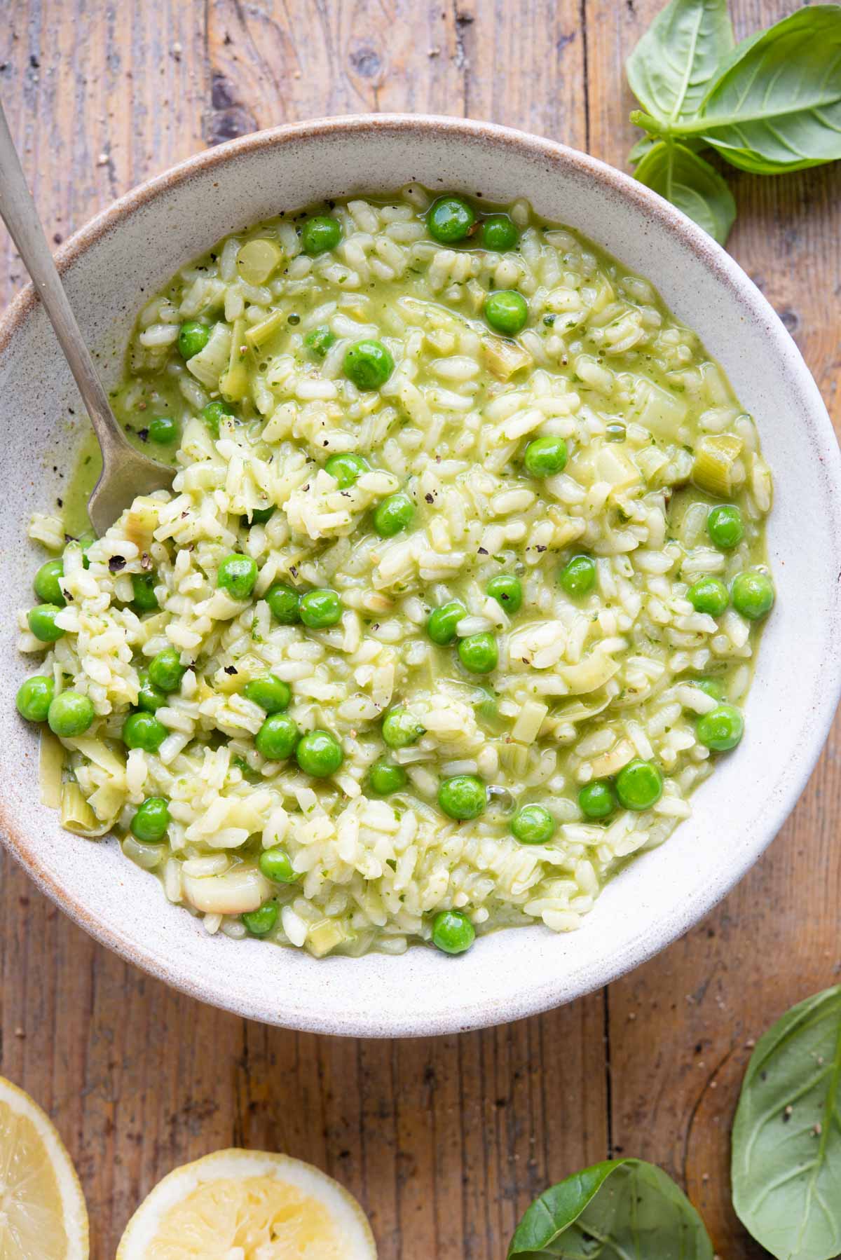 An overhead shot of a bowl of risotto made with peas and pesto on a wooden background with basil and lemons at the side