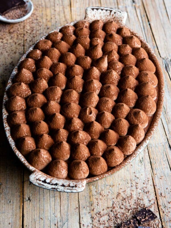 Chocolate tiramisu in an oval dish sitting on a rustic wooden background with pieces of chocolate at the side