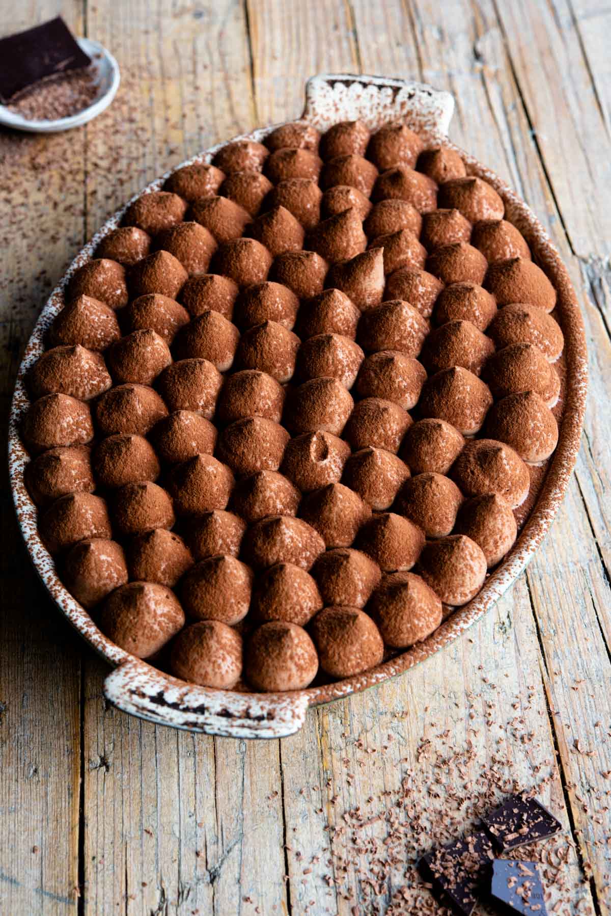 Chocolate tiramisu in an oval dish sitting on a rustic wooden background with pieces of chocolate at the side