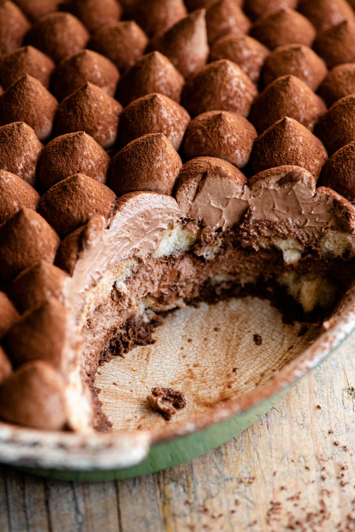 A close up of a chocolate tiramisu in an oval dish with a scoop out revealing layers of chocolate cream and sponge fingers.