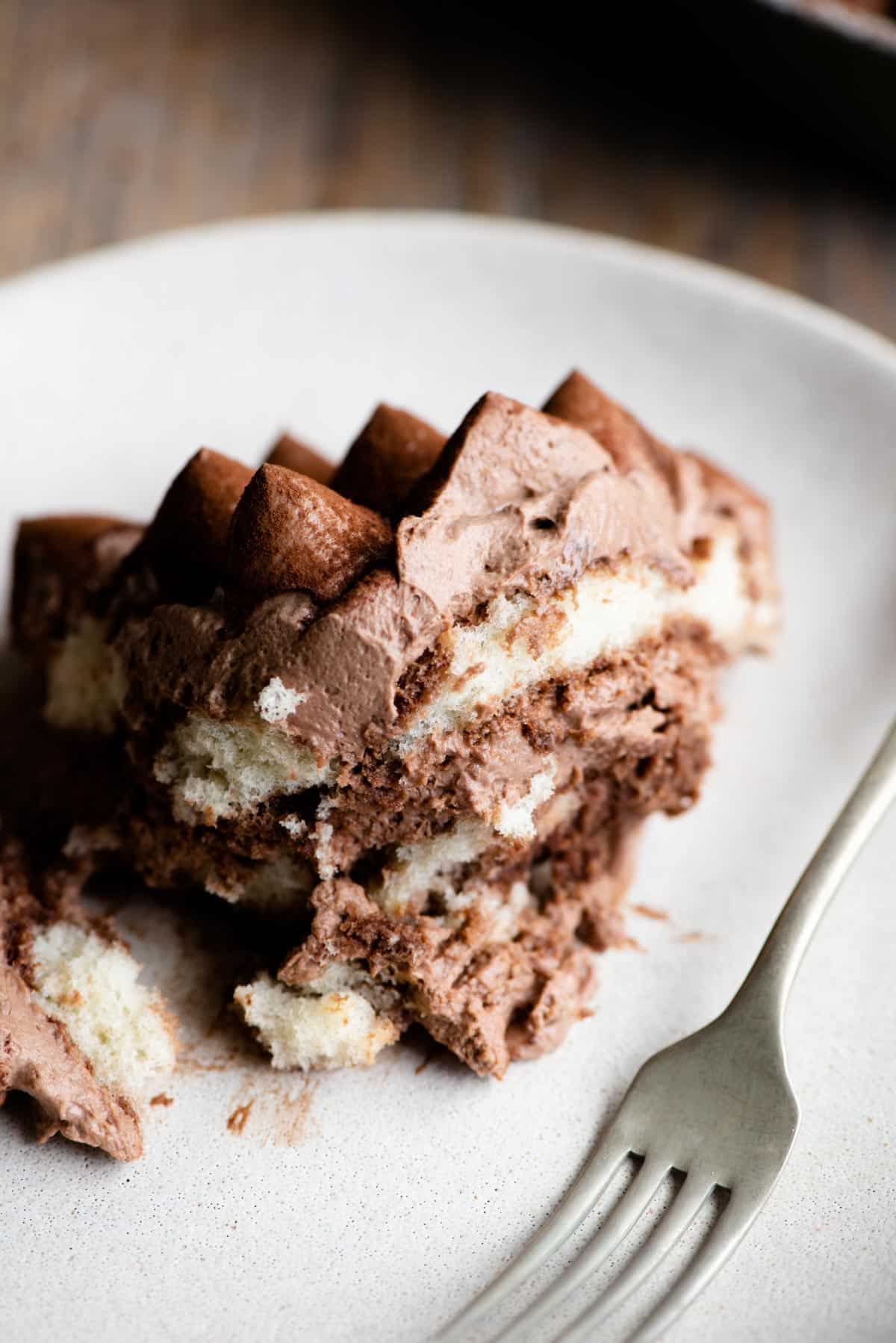 A close up of a piece of chocolate tiramisu on a light coloured plate and a fork at the side.