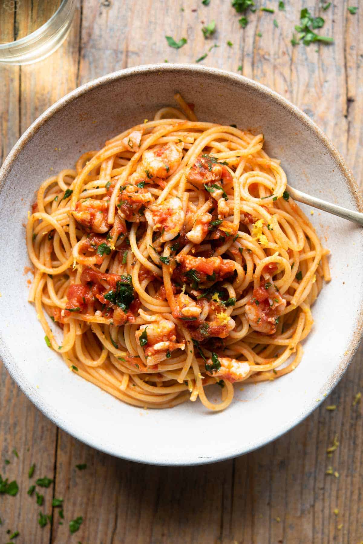 An overhead shot of shrimp spaghetti in a rustic bowl sitting on a wooden surface with a glass of white wine at the side.