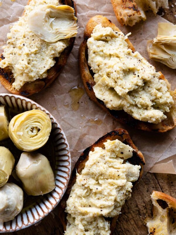 Three slices of artichoke bruschetta sitting on a wooden serving board with a small ramekin of artichokes beside it.