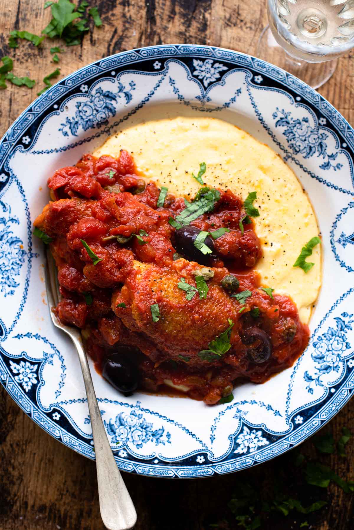 Chicken in Puttanesca sauce in a bowl with polenta sitting on a wooden surface.