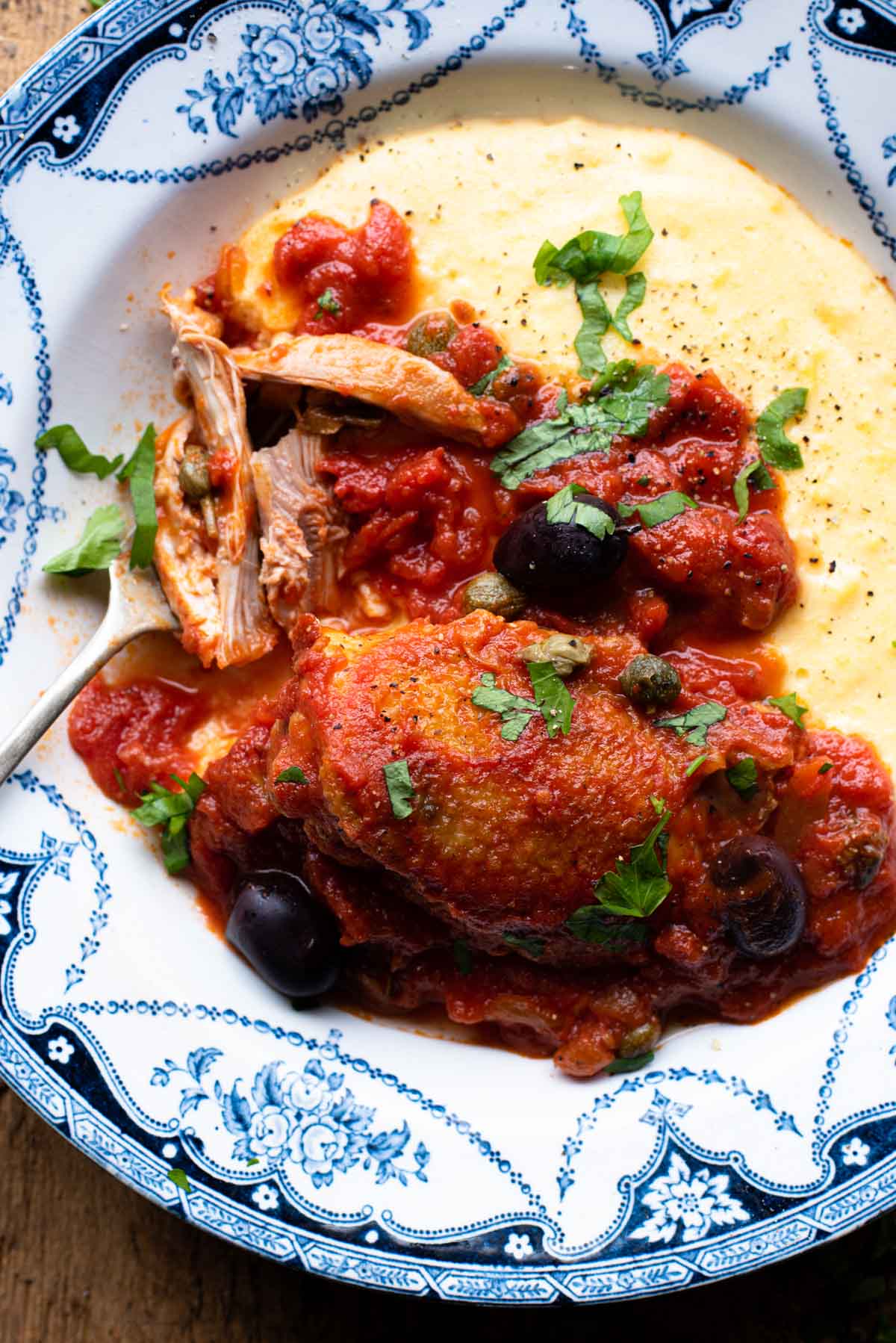 Chicken in Puttanesca sauce in a bowl with polenta sitting on a wooden surface.