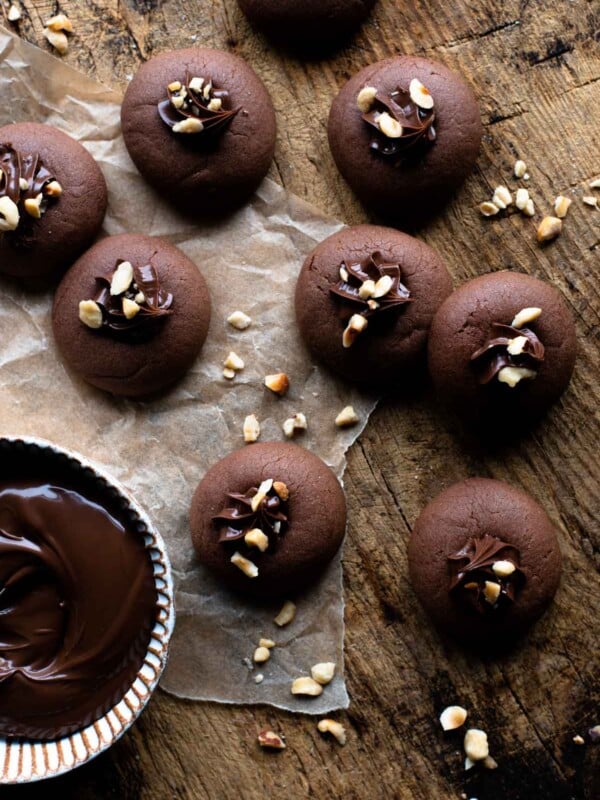 An overhead shot of Nutella cookies on a wooden surface with a ramekin of Nutella at the side and chopped hazelnuts.