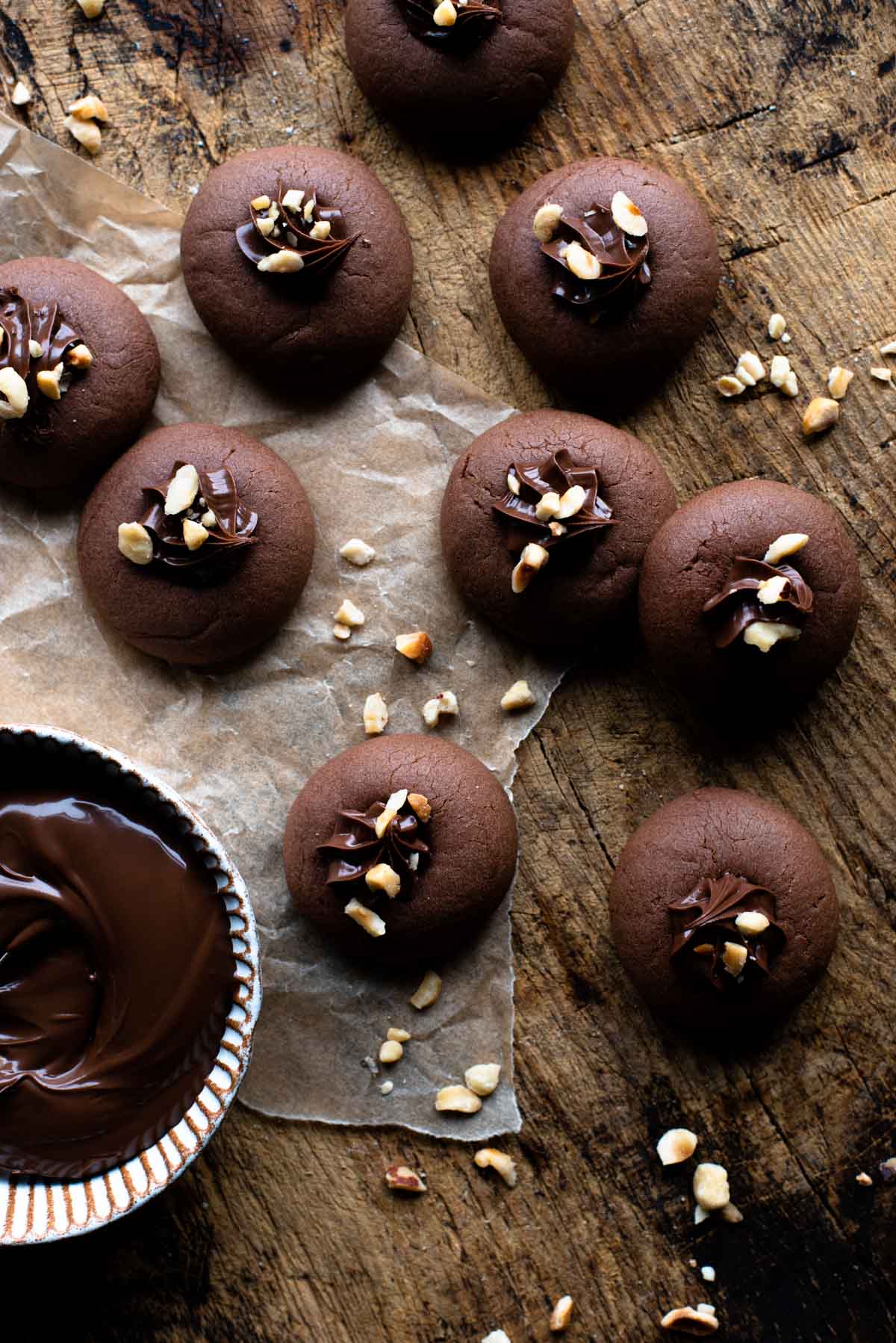 An overhead shot of Nutella cookies on a wooden surface with a ramekin of Nutella at the side and chopped hazelnuts.