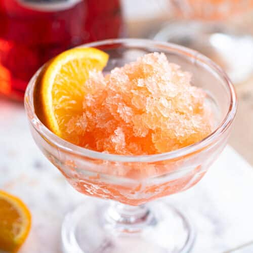 Aperol granita in a small glass bowl with a slice of orange at the side sitting on a marble board.