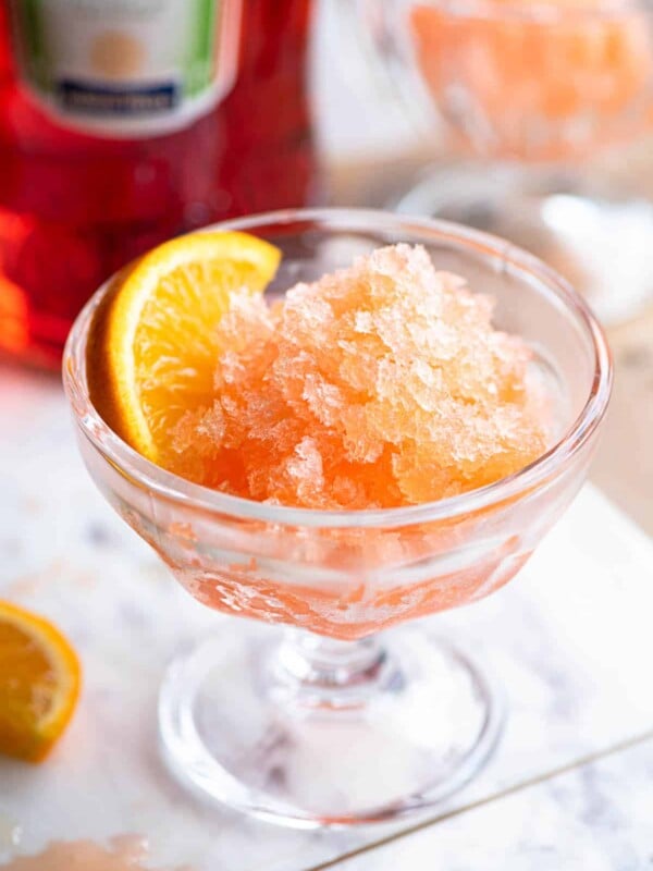 Aperol granita in a small glass bowl with a slice of orange at the side sitting on a marble board.