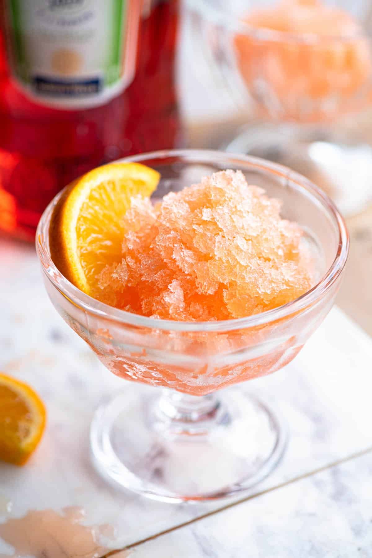 Aperol granita in a small glass bowl with a slice of orange at the side sitting on a marble board.