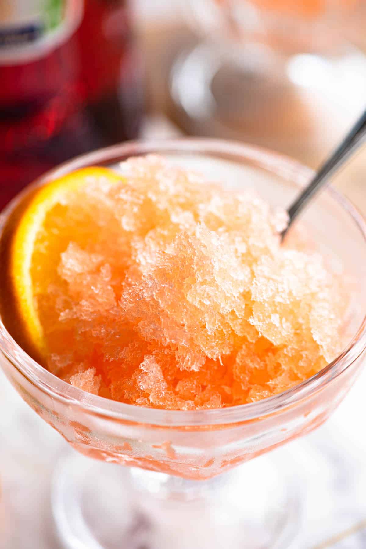 A close up of Aperol granita in a small glass bowl with a spoon and slice of orange at the side.