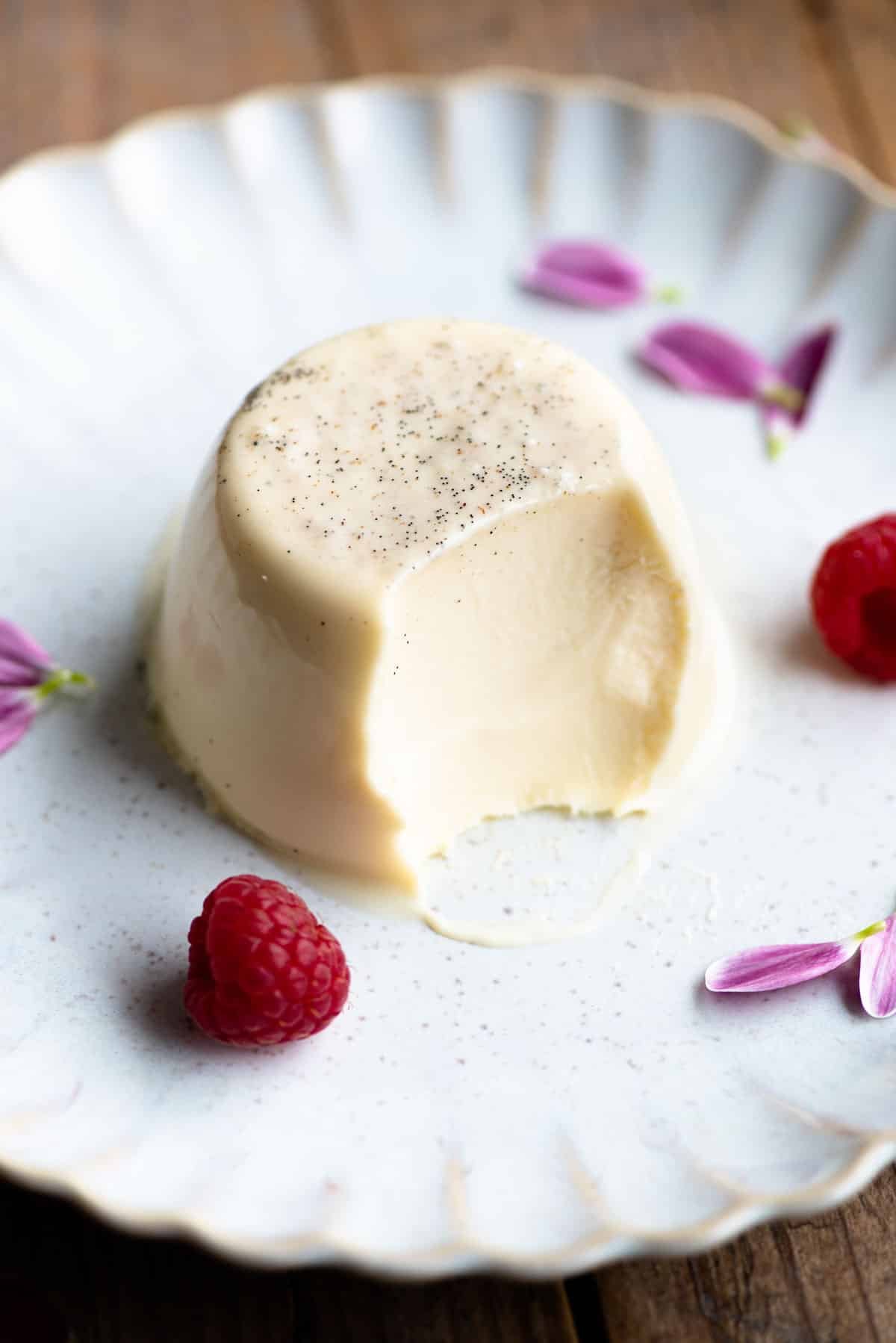 A vanilla panna cotta on a plate with a bite out and raspberries at the side.