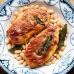 A cropped square image of chicken saltimbocca in a blue bowl on top of cannellini beans.