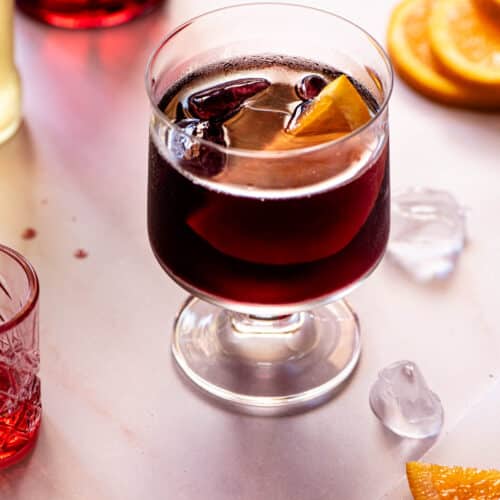 A red wine spritz (prampolini) on a light background with slices of orange, ice and campari at the side.