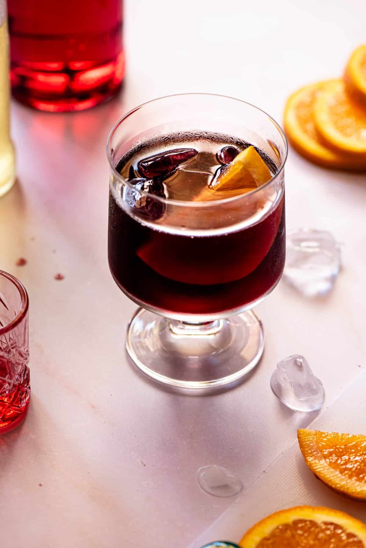 A red wine spritz (prampolini) on a light background with slices of orange, ice and campari at the side.