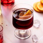 A square image of an Italian red wine spritz on a light work surface with ice and oranges around.