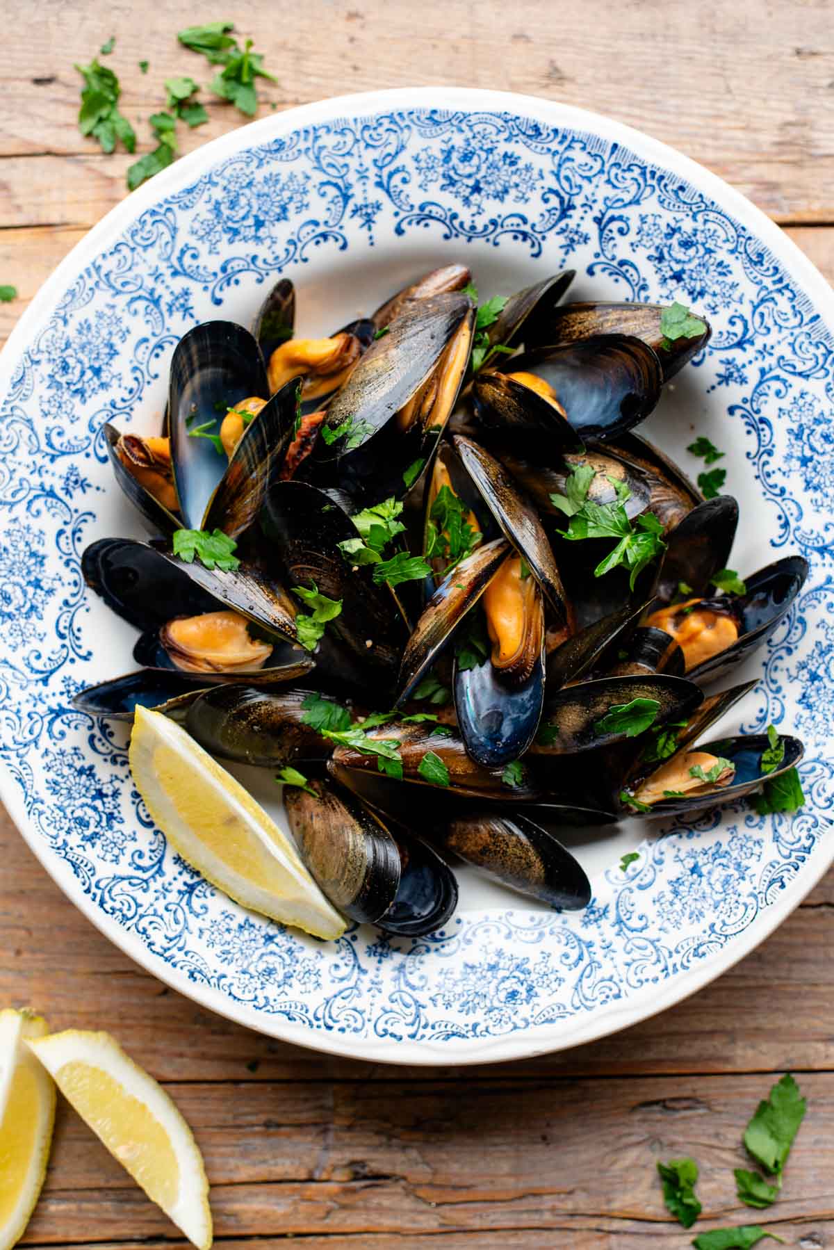 An overhead shot of mussels in a blue bowl with lemon wedges.