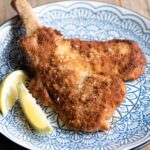 A cropped square image of Veal Milanese on a blue plate with a lemon wedge at the side.