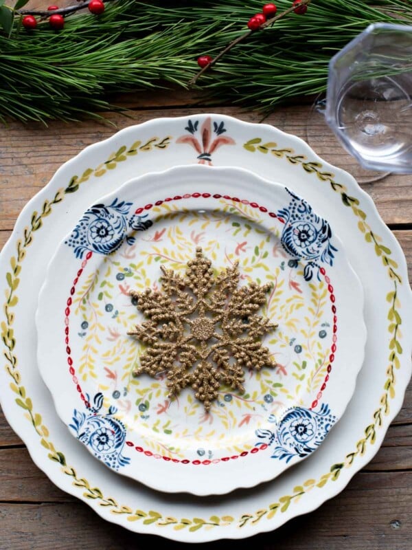 A Christmas table setting with Christmas plates on a wooden table and foliage at the top if the table.