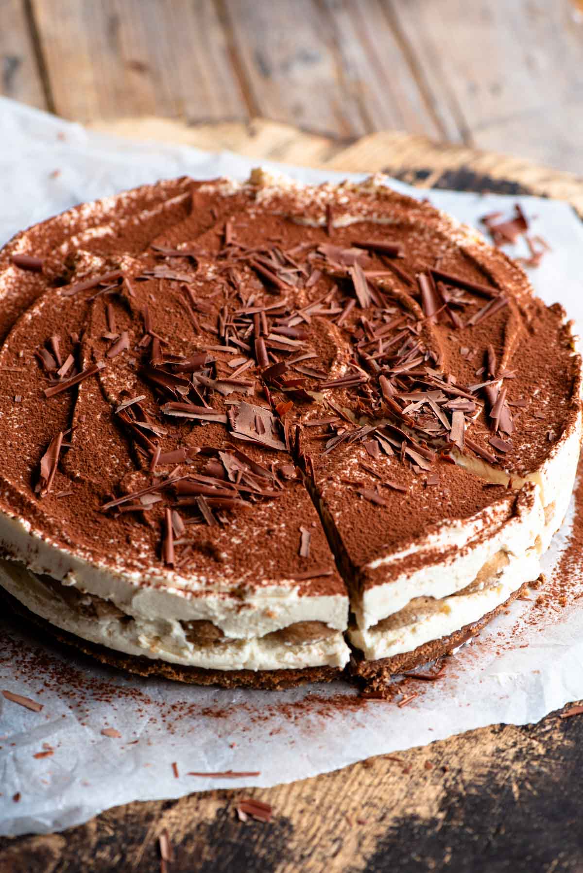 A whole tiramisu cheesecake sitting on a wooden board and parchment paper.