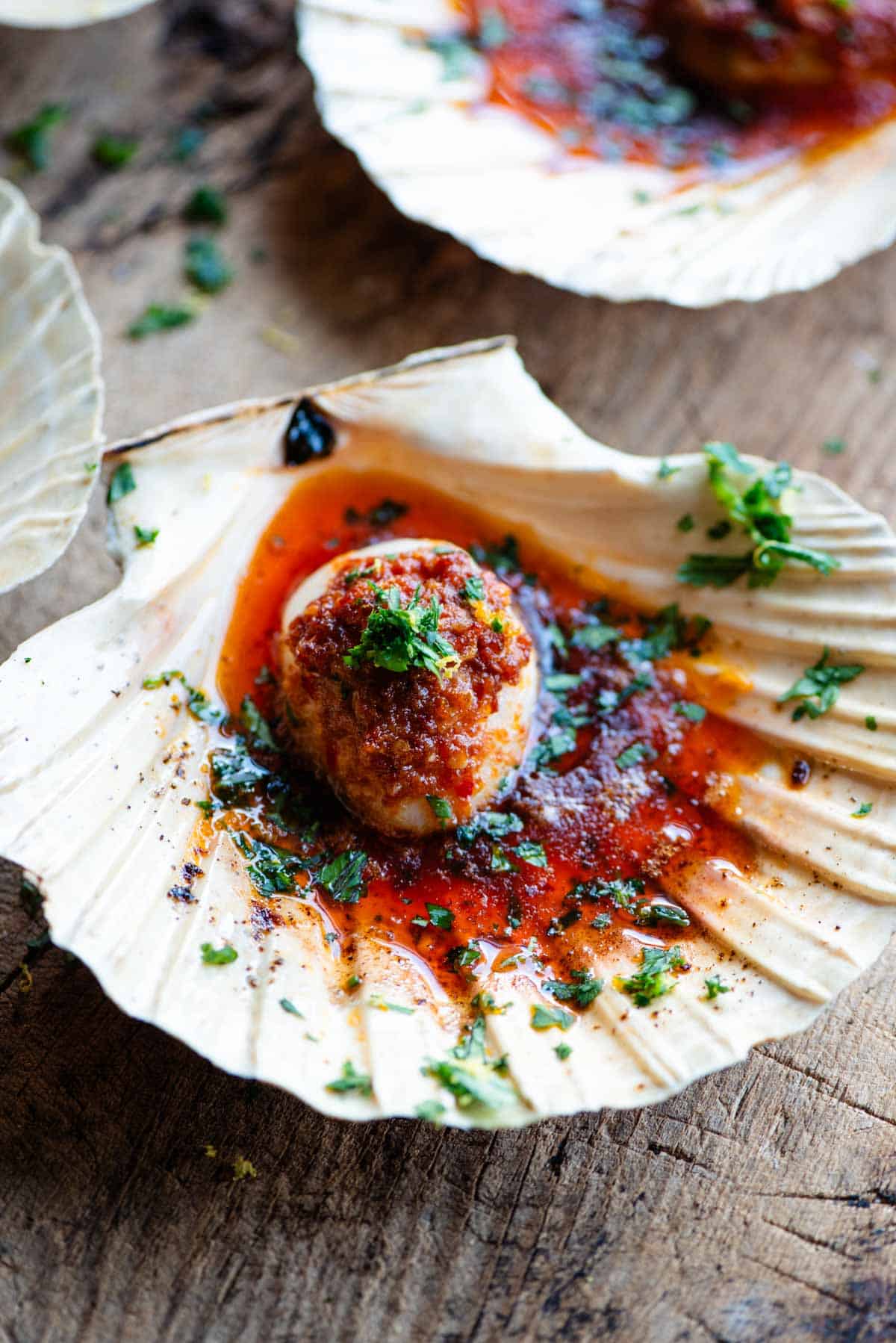A close up side shot of a scallop in its shell with Nduja garlic butter on top and garnished with parsley.
