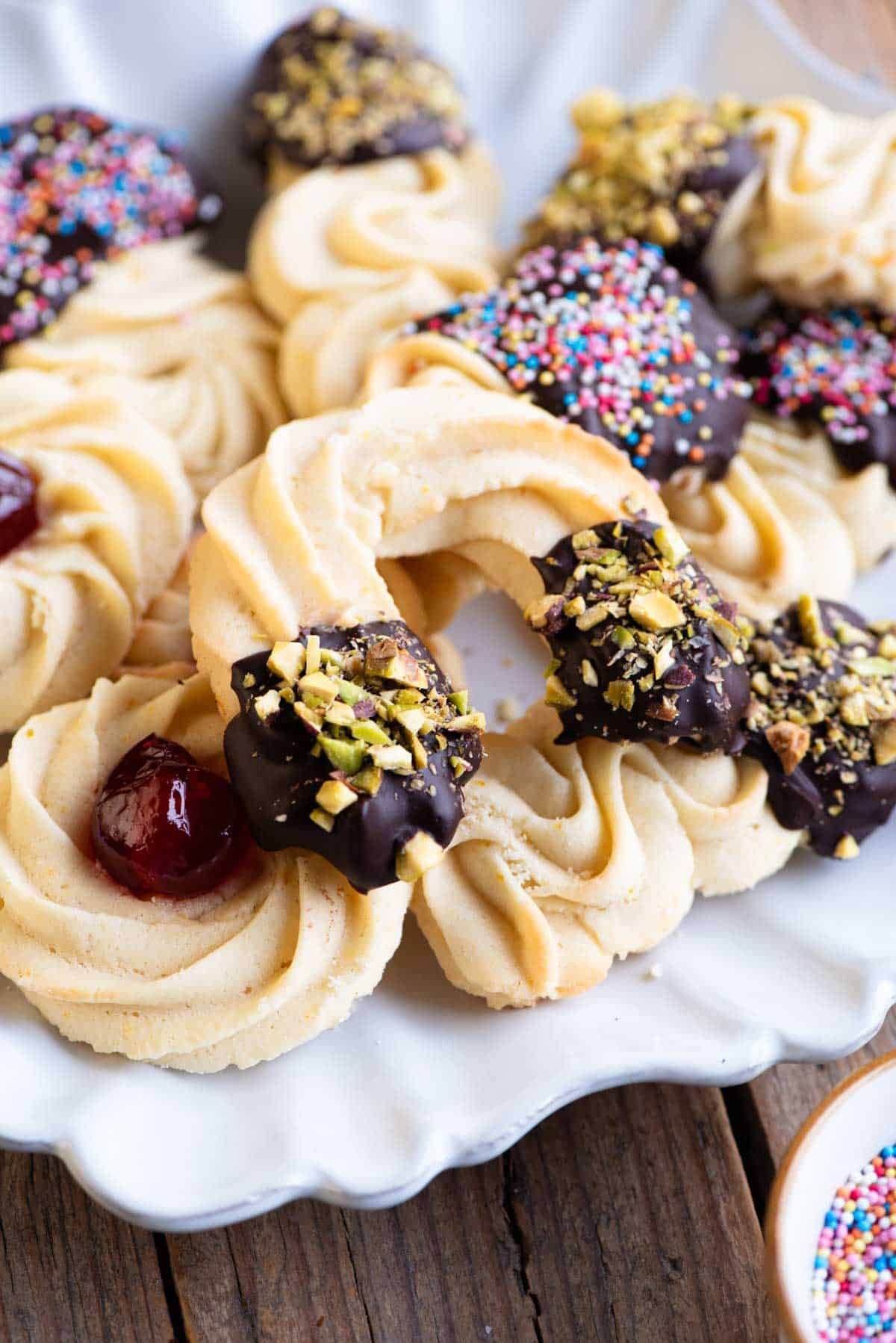 A close up of Italian butter cookies on a plate some half dipped in chocolate and sprinkled with pistachios.