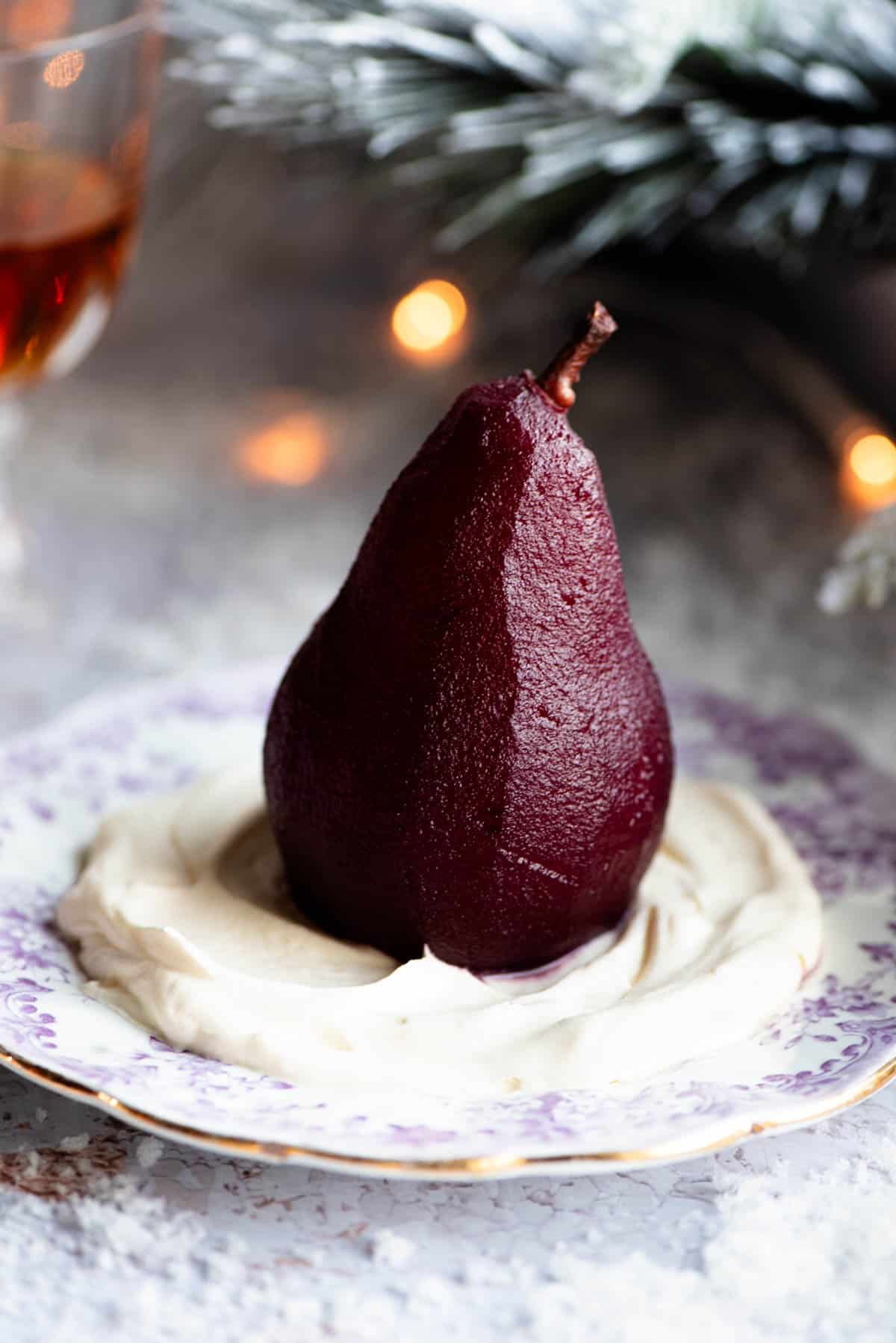 A red wine poached pear sitting on a bed of Amaretto whipped cream on a small plate, the background is Christmassy.