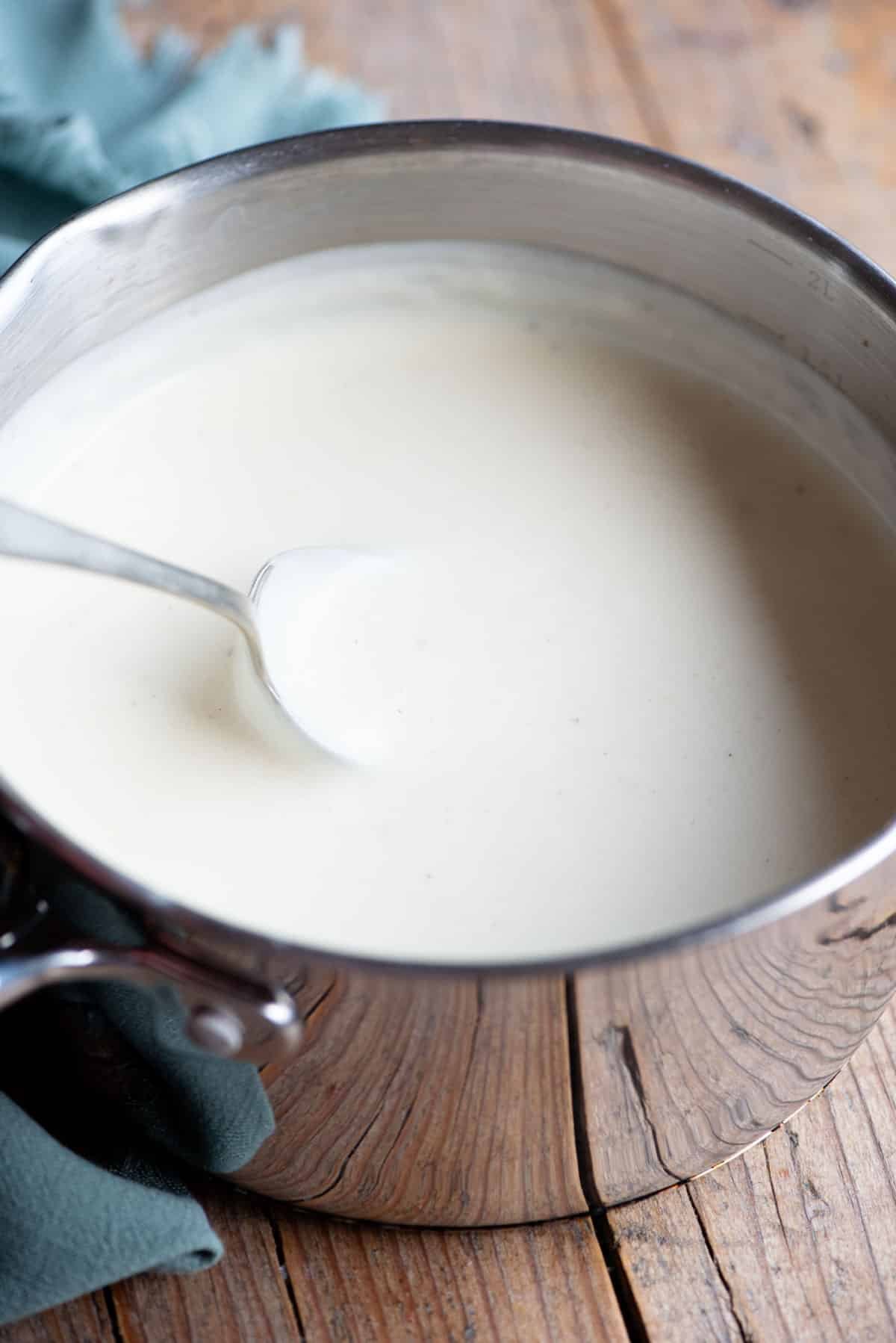 A close up of bechamel sauce in a pot with a spoon. The background is wooden.