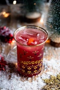 A close up of a Cranberry and Prosecco cocktail in a glass with snow and Christmas decorations all around.