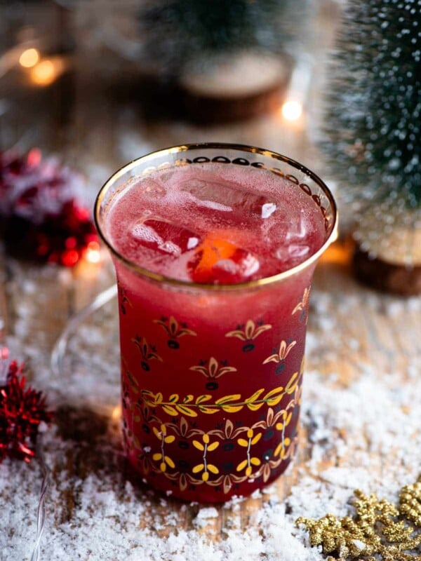A close up of a Cranberry and Prosecco cocktail in a glass with snow and Christmas decorations all around.
