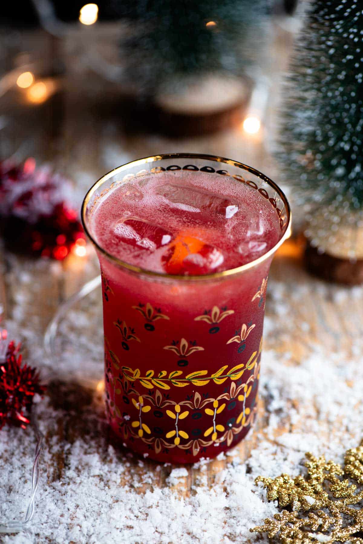 A close up of a Cranberry and Prosecco cocktail in a glass with snow and Christmas decorations all around.