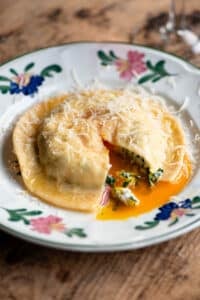 A side shot of one large egg yolk ravioli on a patterned plate. The ravioli is cut open and egg yolk is running out.