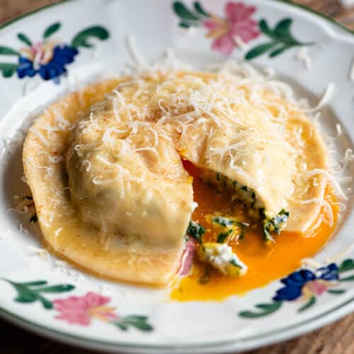 A side shot of one large egg yolk ravioli on a patterned plate. The ravioli is cut open and egg yolk is running out.