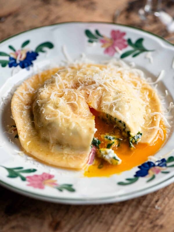 A side shot of one large egg yolk ravioli on a patterned plate. The ravioli is cut open and egg yolk is running out.