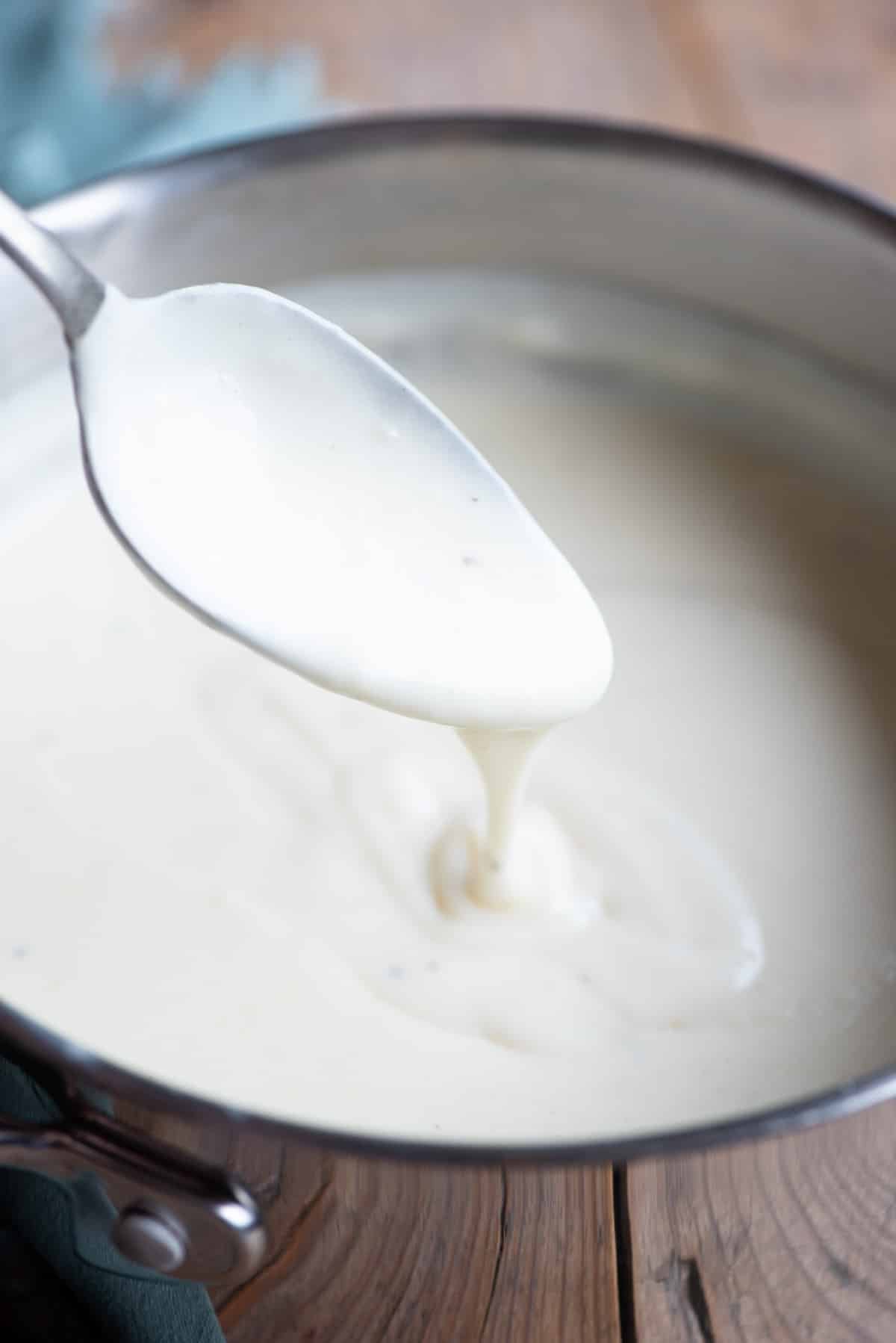 A close up of Italian Besciamella sauce in a pot with some dripping off a spoon.