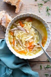 An overhead shot of a bowl of lemon chicken orzo soup on a wooden surface with crusty bread at the side.