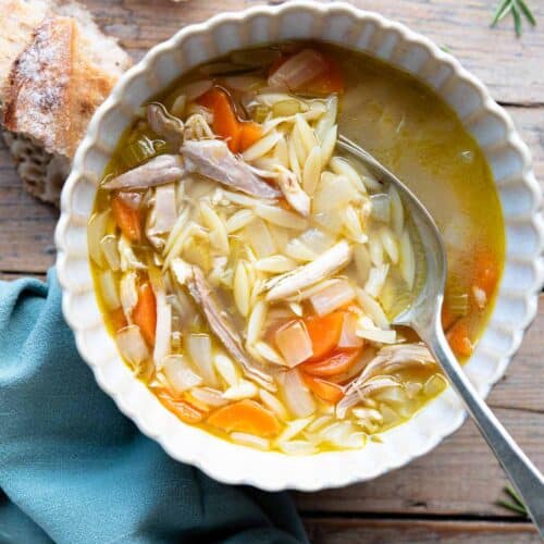 An overhead shot of a bowl of lemon chicken orzo soup on a wooden surface with crusty bread at the side.