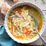 An overhead shot of lemon chicken orzo soup on a bowl. The background is wooden with crusty bread at the side.